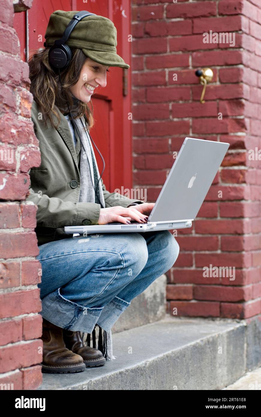 Hip woman relaxes infront of red door while working on laptop and listening to music. Stock Photo