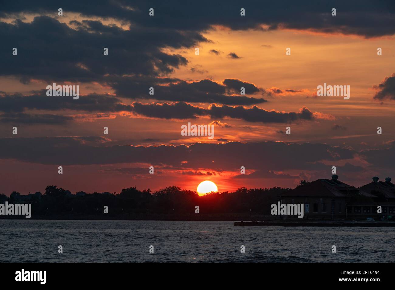The setting sun behind the silhouetted trees and water creates a dramatic cloudscape. Stock Photo