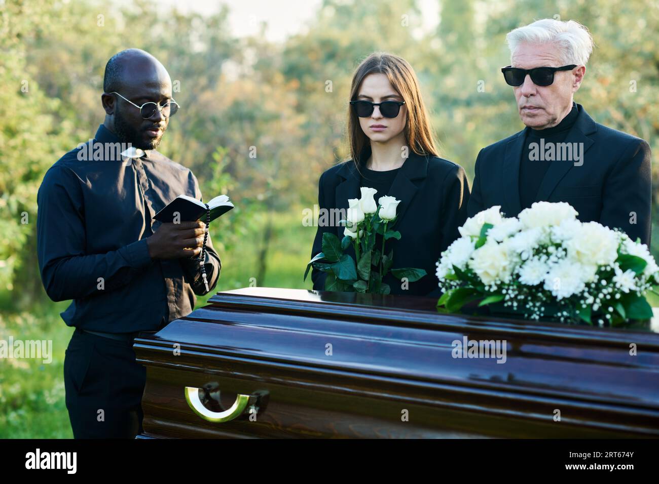 Mature man and young woman in mourning clothes and sunglasses standing by coffin while priest