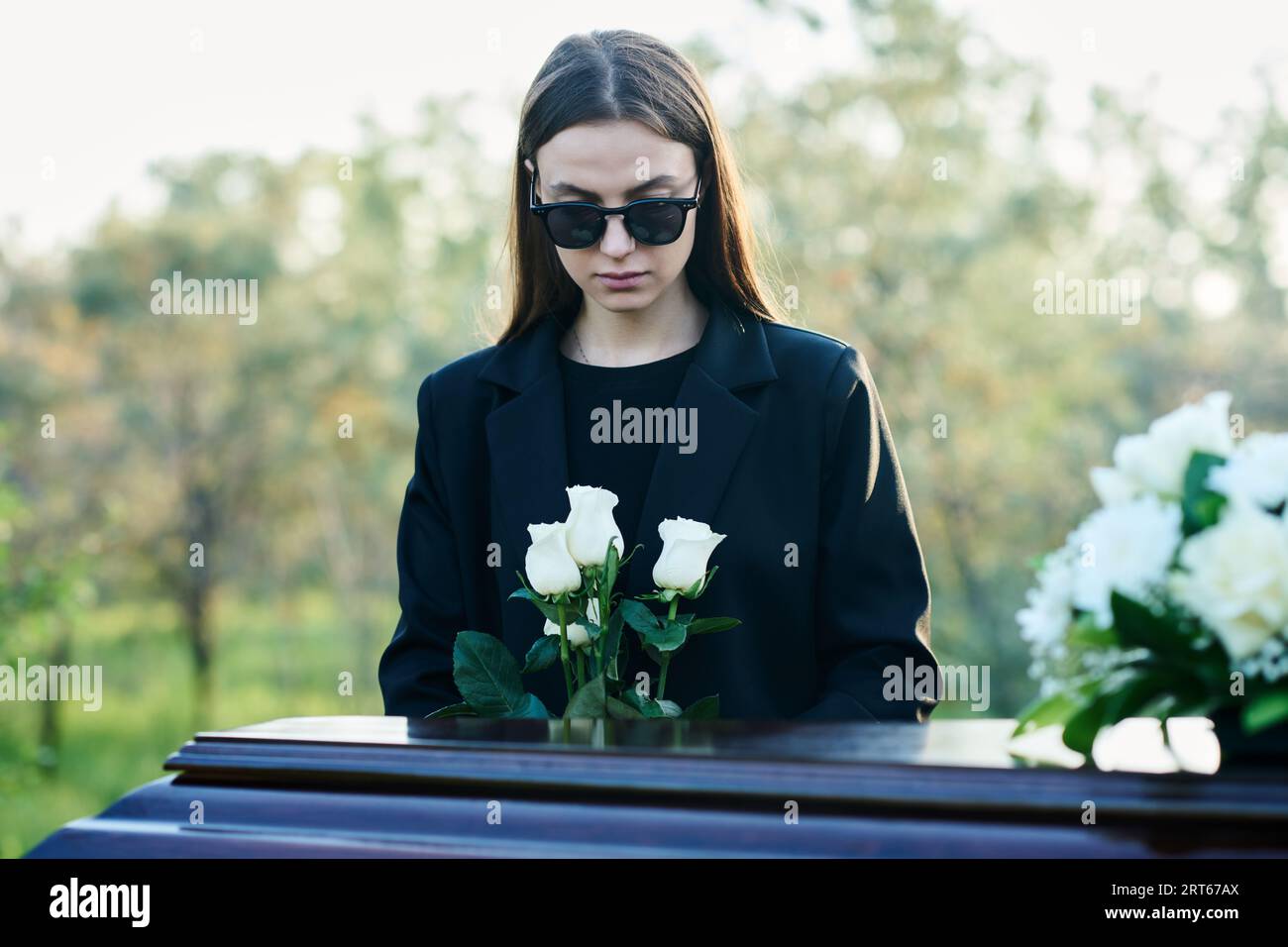 Young widow with bunch of fresh white roses standing by coffin with