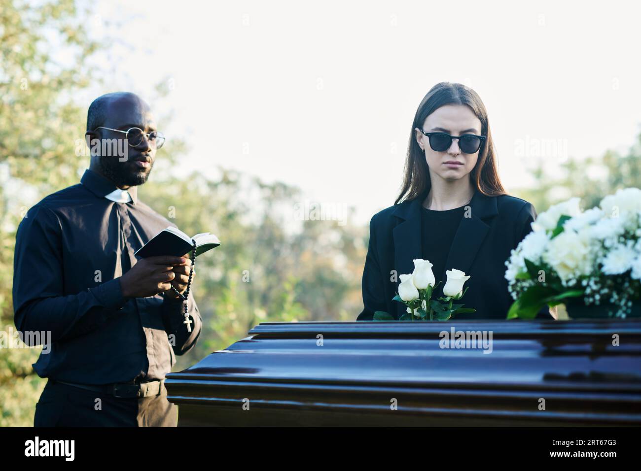 Young inconsolable widow in mourning attire and sunglasses holding bunch of white roses while standing by coffin next to priest with Bible Stock Photo