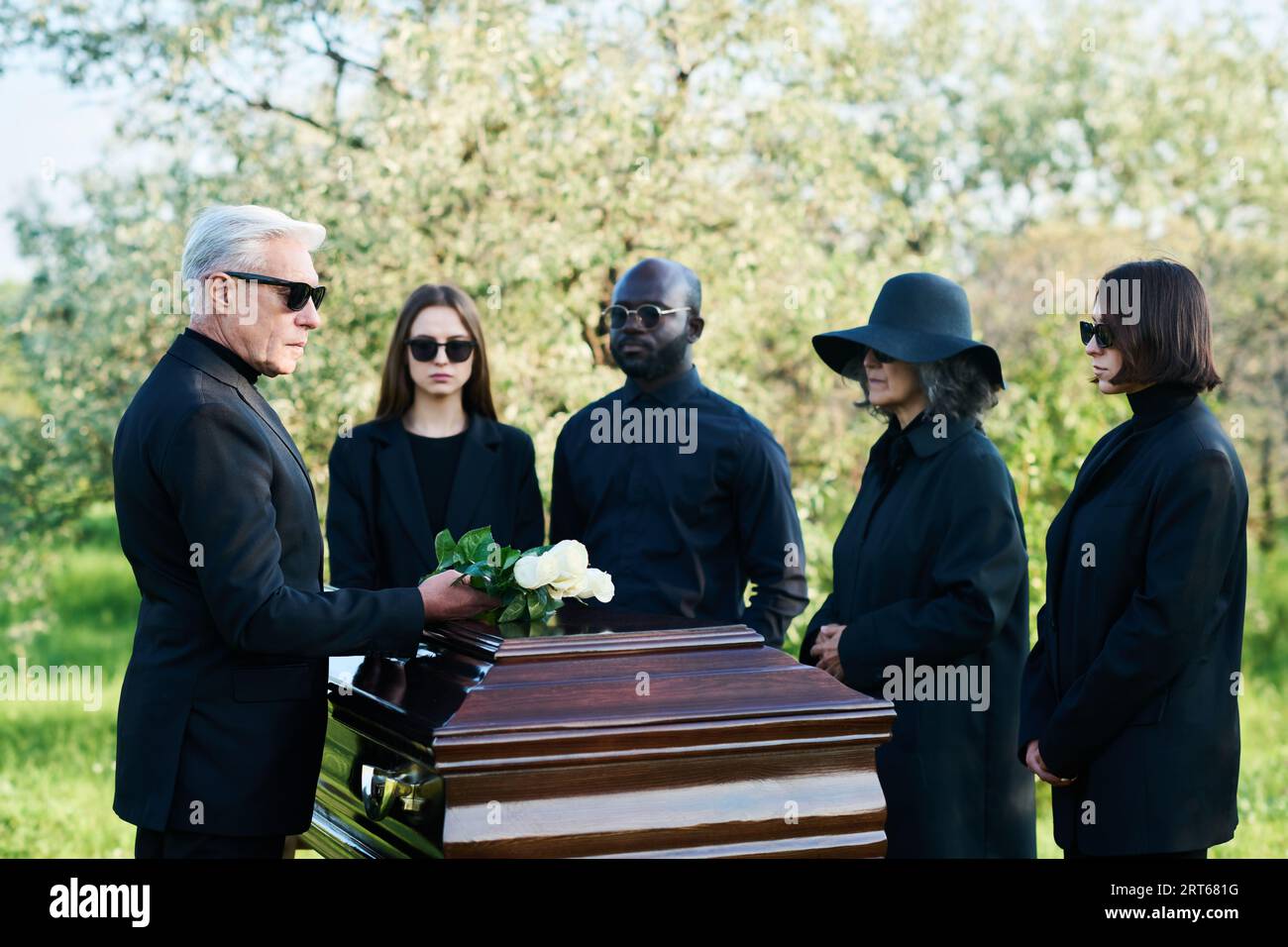 Mature man in black suit and sunglasses putting bunch of white roses on top of closed lid of coffin while standing among other mourning people Stock Photo