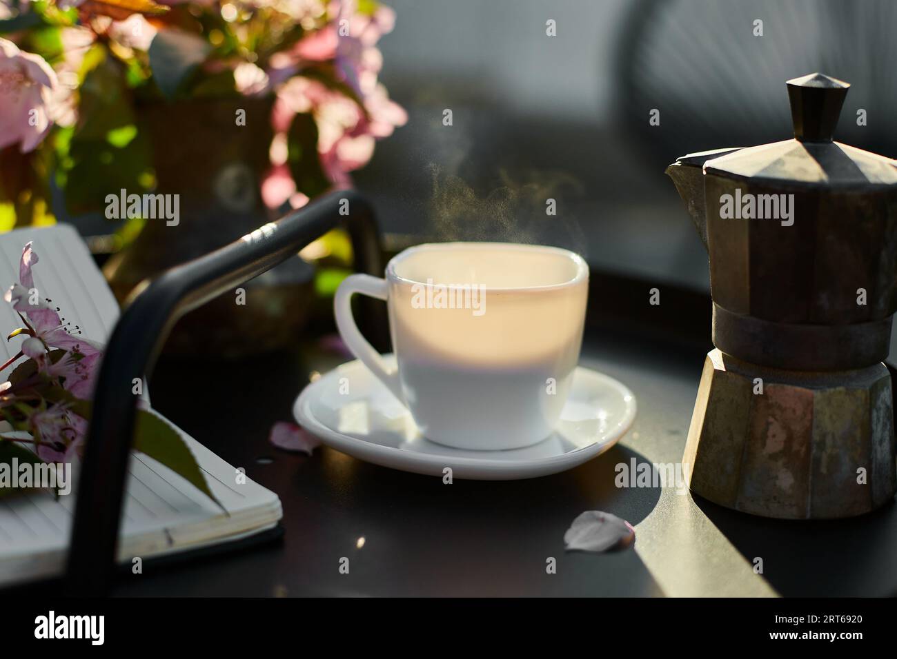 https://c8.alamy.com/comp/2RT6920/cup-of-coffee-with-steam-flowers-and-notebook-on-the-small-black-table-on-terrace-at-home-in-a-sunny-day-outdoor-workspace-summer-relaxation-coffee-2RT6920.jpg
