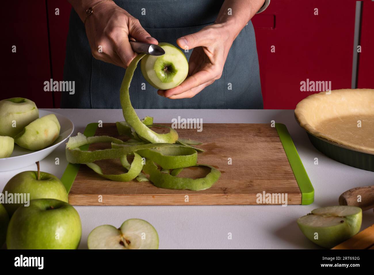 Woman is peeling apples to prepare apple pie in the kitchen. Authentic female hands peel an apple with a knife. Cooking apple pie process, step by ste Stock Photo