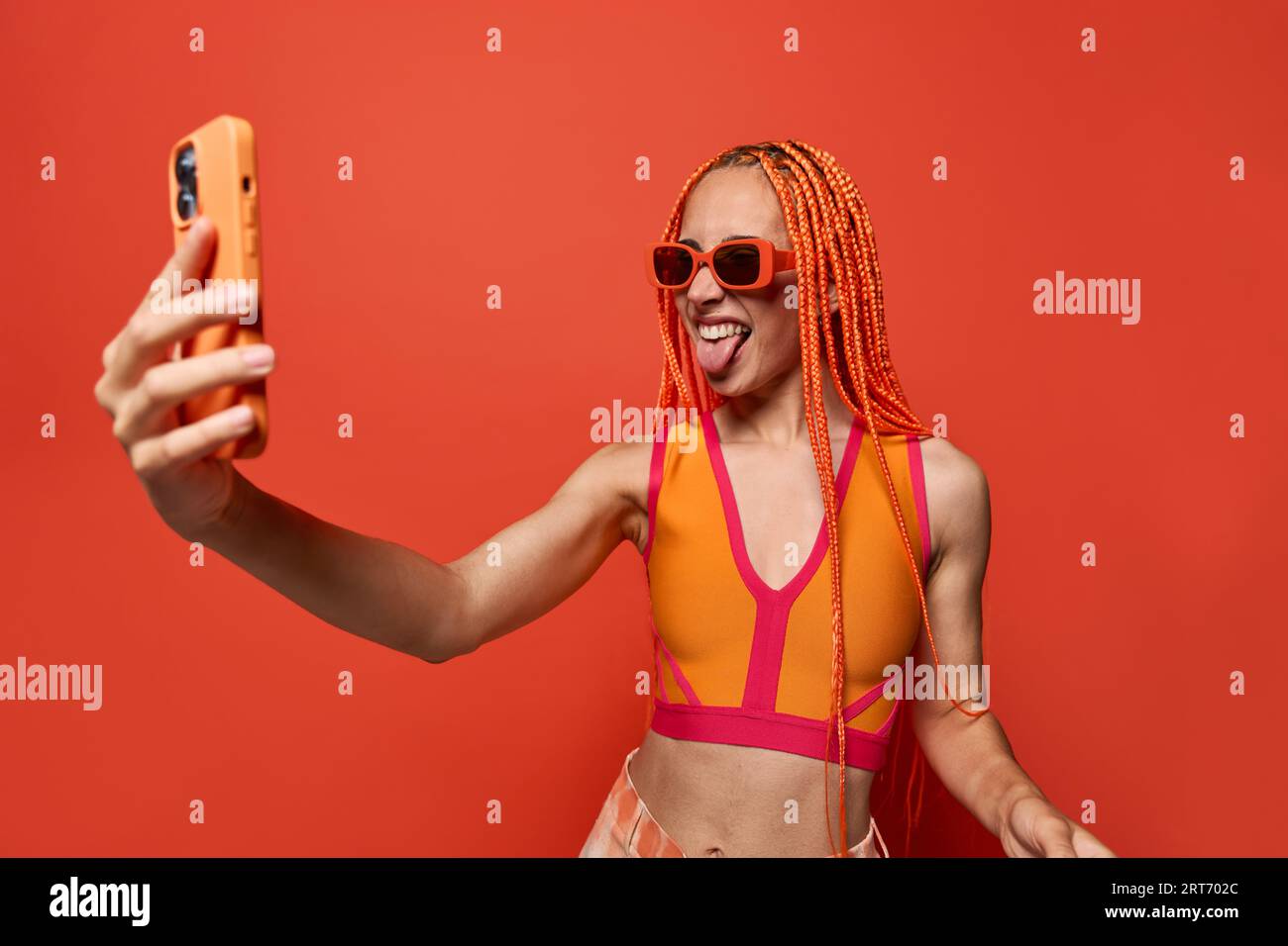 Young stylish woman with Afro braids in sunglasses and colorful top taking selfie on cellphone against red background Stock Photo