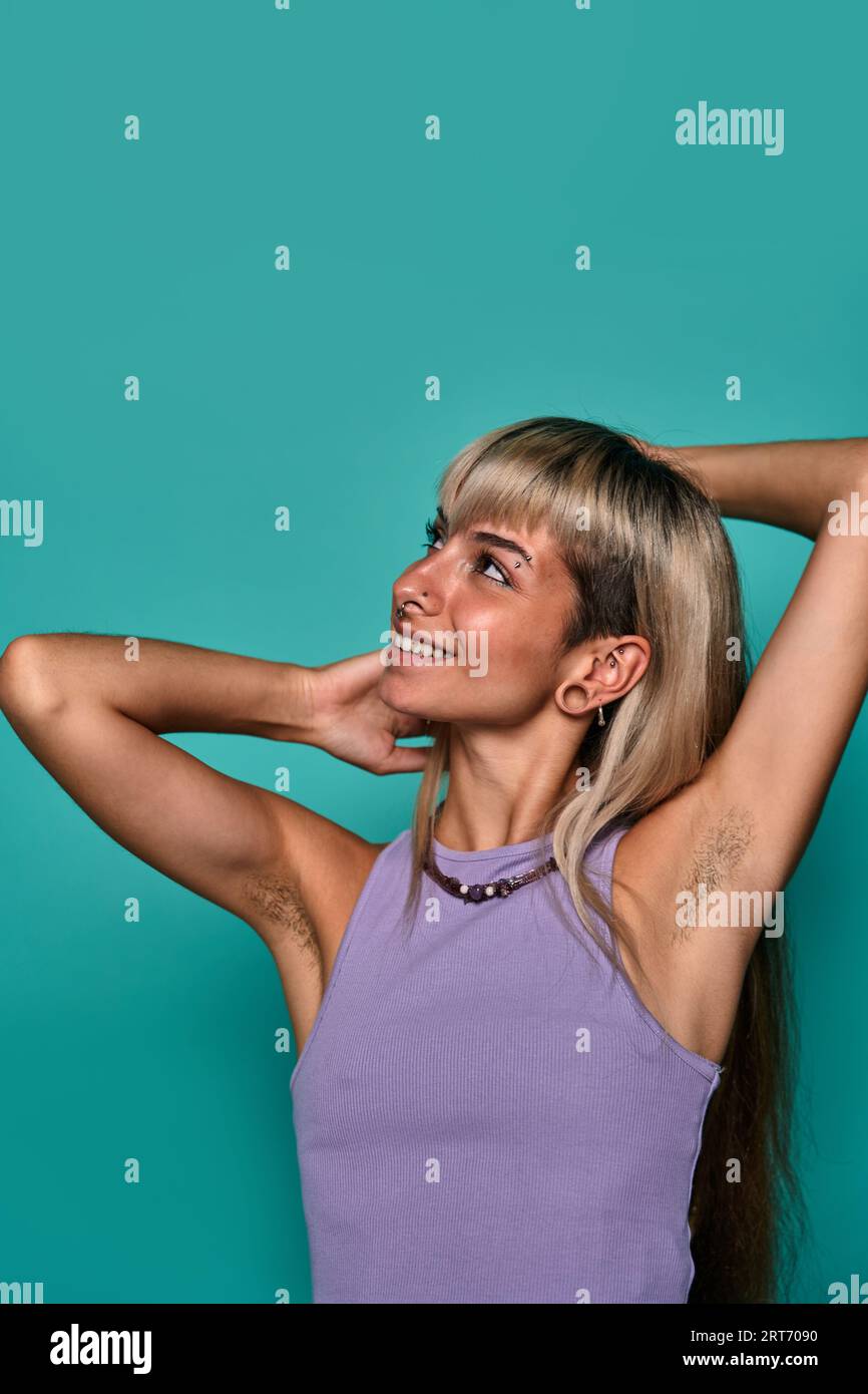 Young female with blond hair and piercing putting arms behind head while showing hairy armpit against turquoise background body positive concept Stock Photo