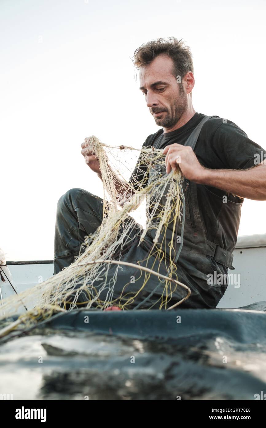 Focused male sitting and untangling fishing net on wet boat deck with