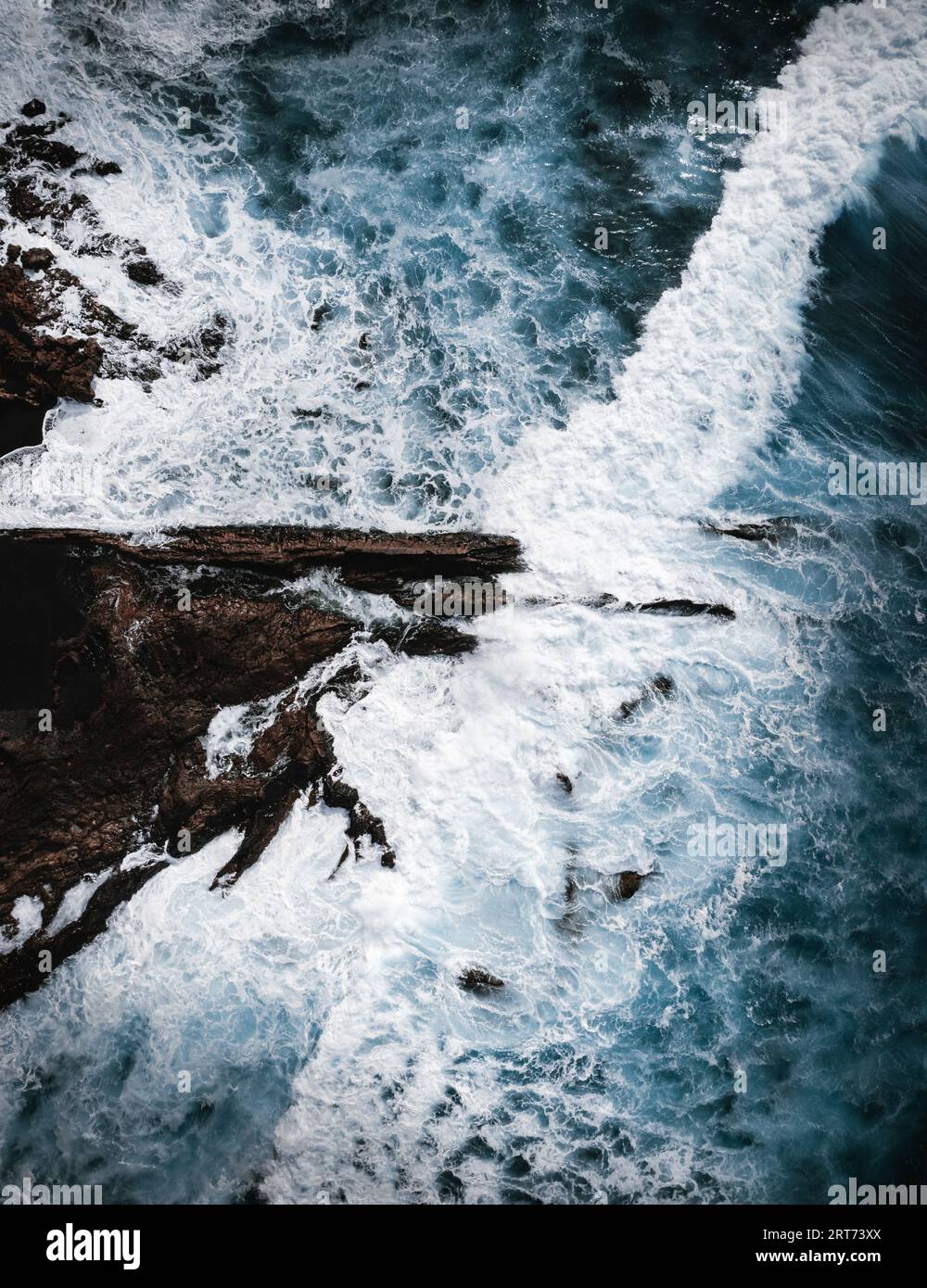 Vertical Aerial photo of strong and powerful ocean with huge waves. Rocks and part of cliff inside the stormy blue ocean - top view. Dark and moody ph Stock Photo