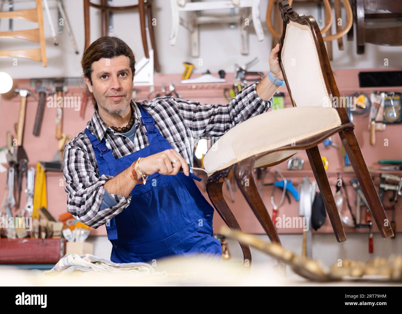 Middle-aged male restorer removes old cloth seat covering from antique wooden chair. Stock Photo