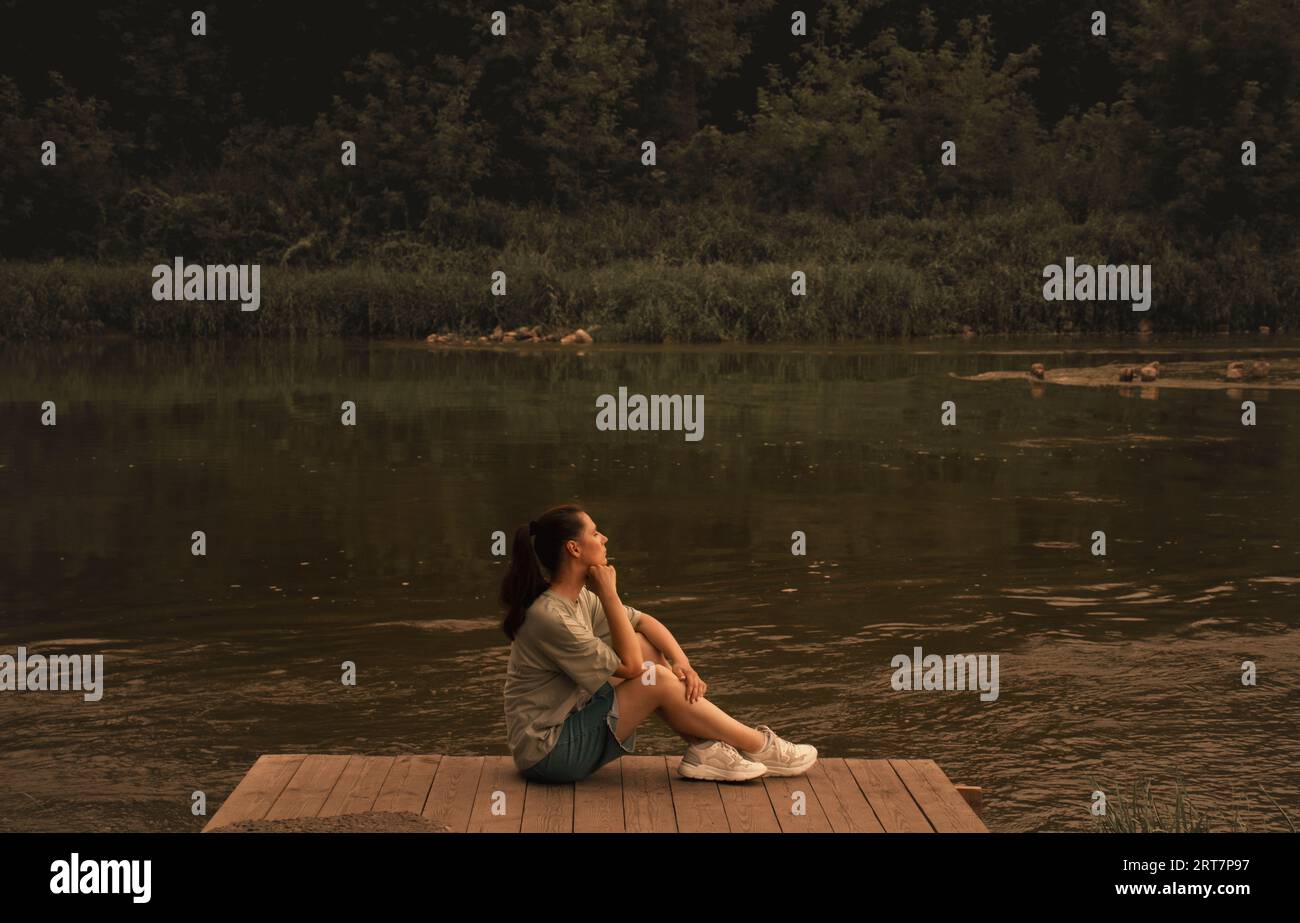 young woman sits calmly on the river bank and watches the sunset Stock Photo