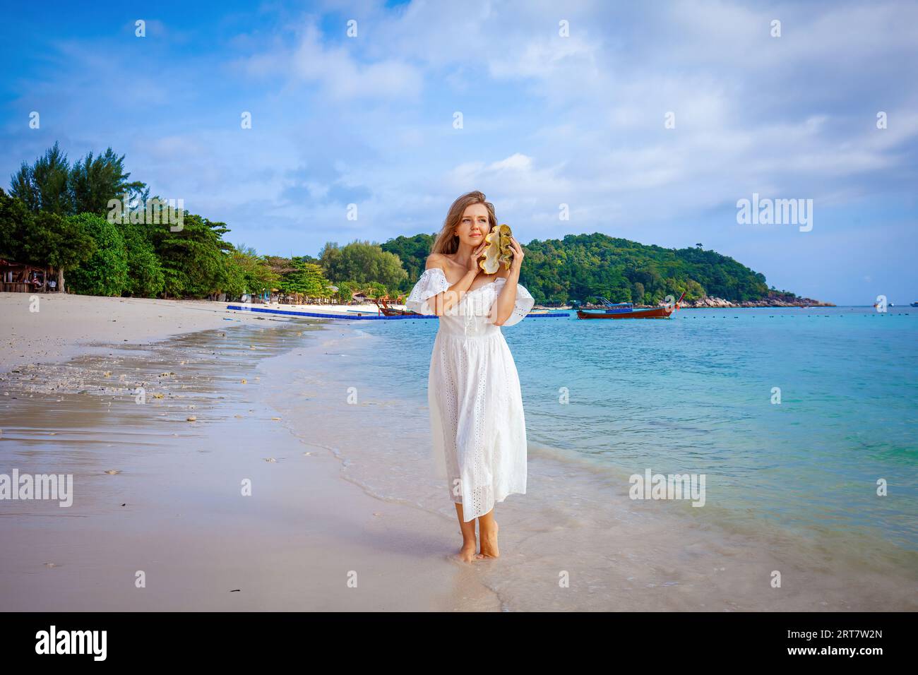 Beautiful woman in white dress strolls on sandy beach with seashell in ...