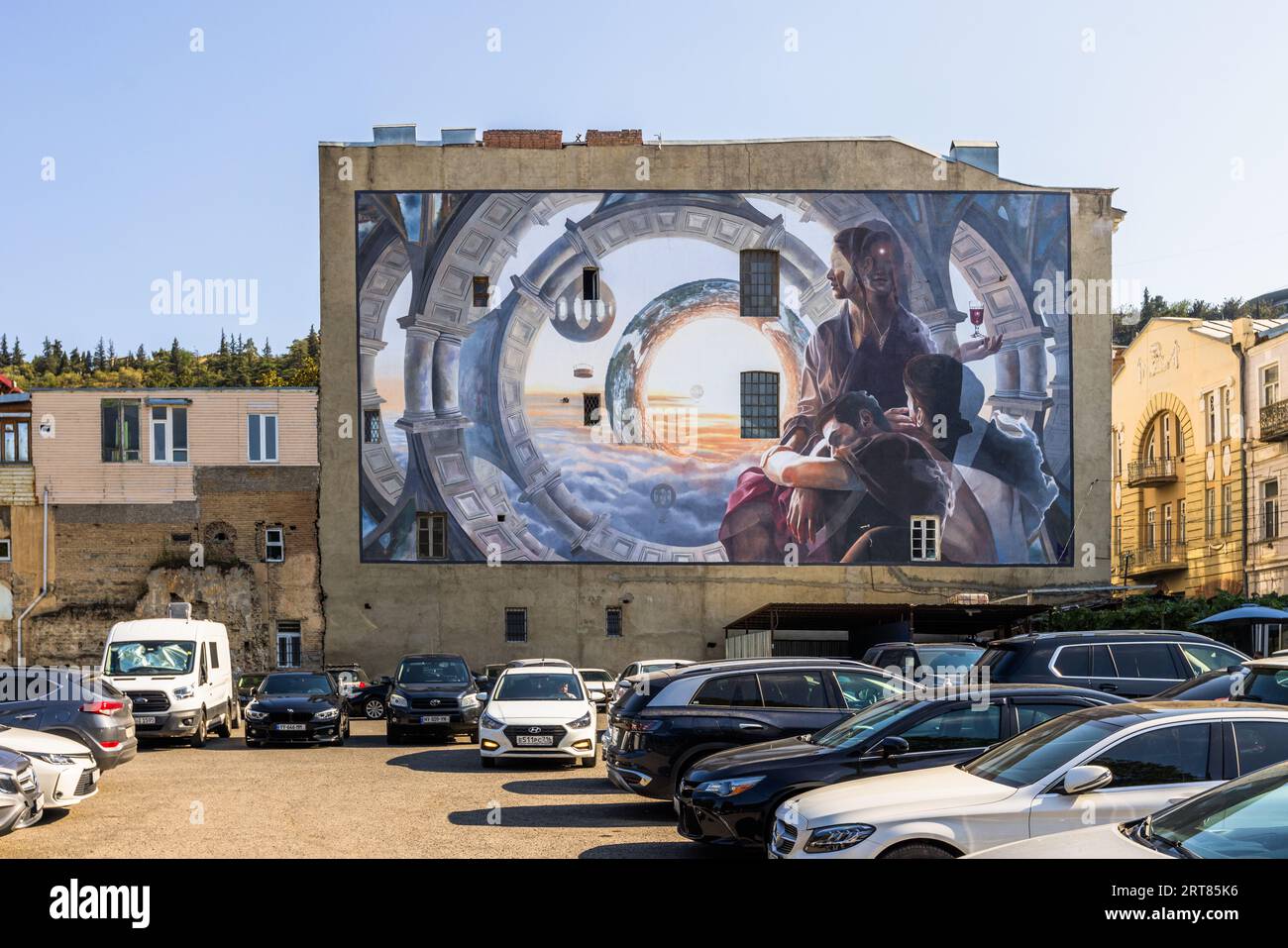 A mural covers the side of a building in Tbilisi, Georgia. The painting shows architectural elements, clouds and people. Cars park in front of the building Stock Photo