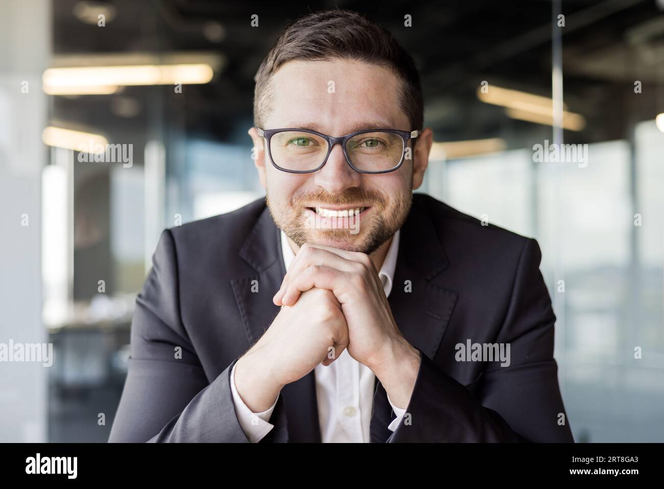 Close-up photo of a young smiling man, businessman, director, sitting in the office in a suit, resting his head on his hands and looking at the camera with a smile. Stock Photo