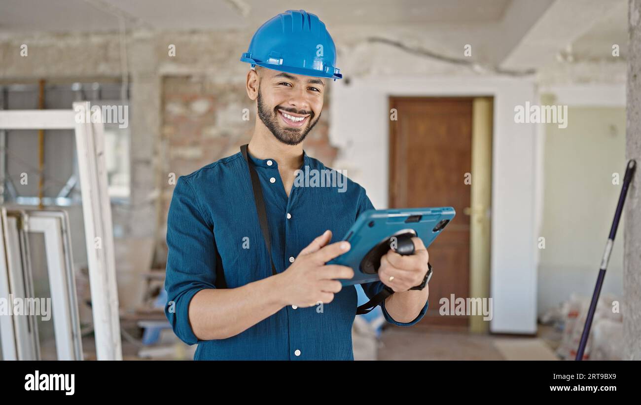 Young hispanic man architect wearing hardhat smiling using tablet at construction site Stock Photo