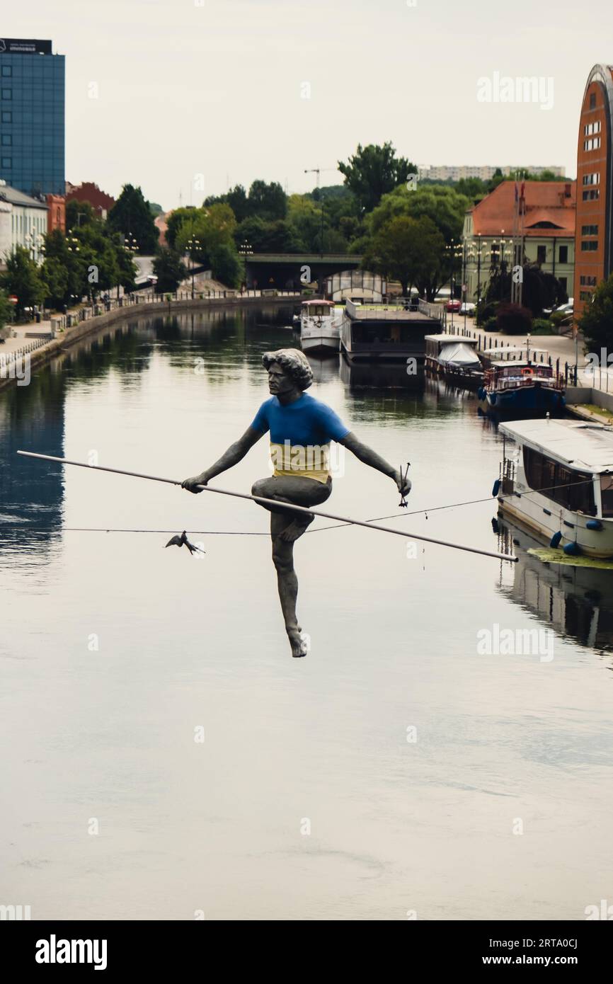 Bydgoszcz, Poland - August 2022 Brda river in Bydgoszcz, Man crossing a river sculpture , of a man balancing on a wire, old granary building, Kuyavian-Pomerania. Old town with reflection in Brda River. Old town view with famous statue in Ukrainian flag on the rope. Stock Photo