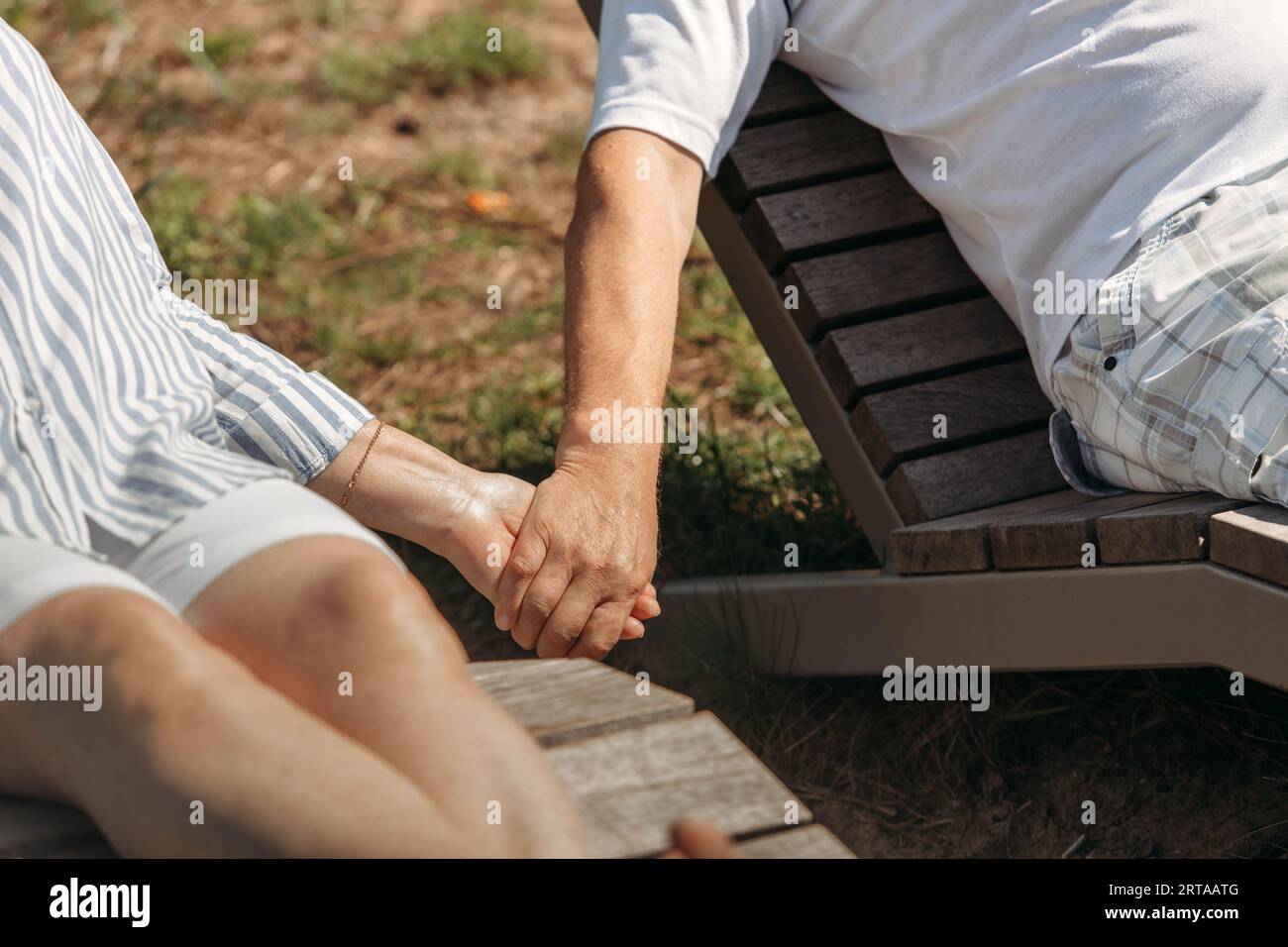 A couple of cute elderly people lie on sunbeds holding hands, basking in the sun. Close-up of hands Stock Photo