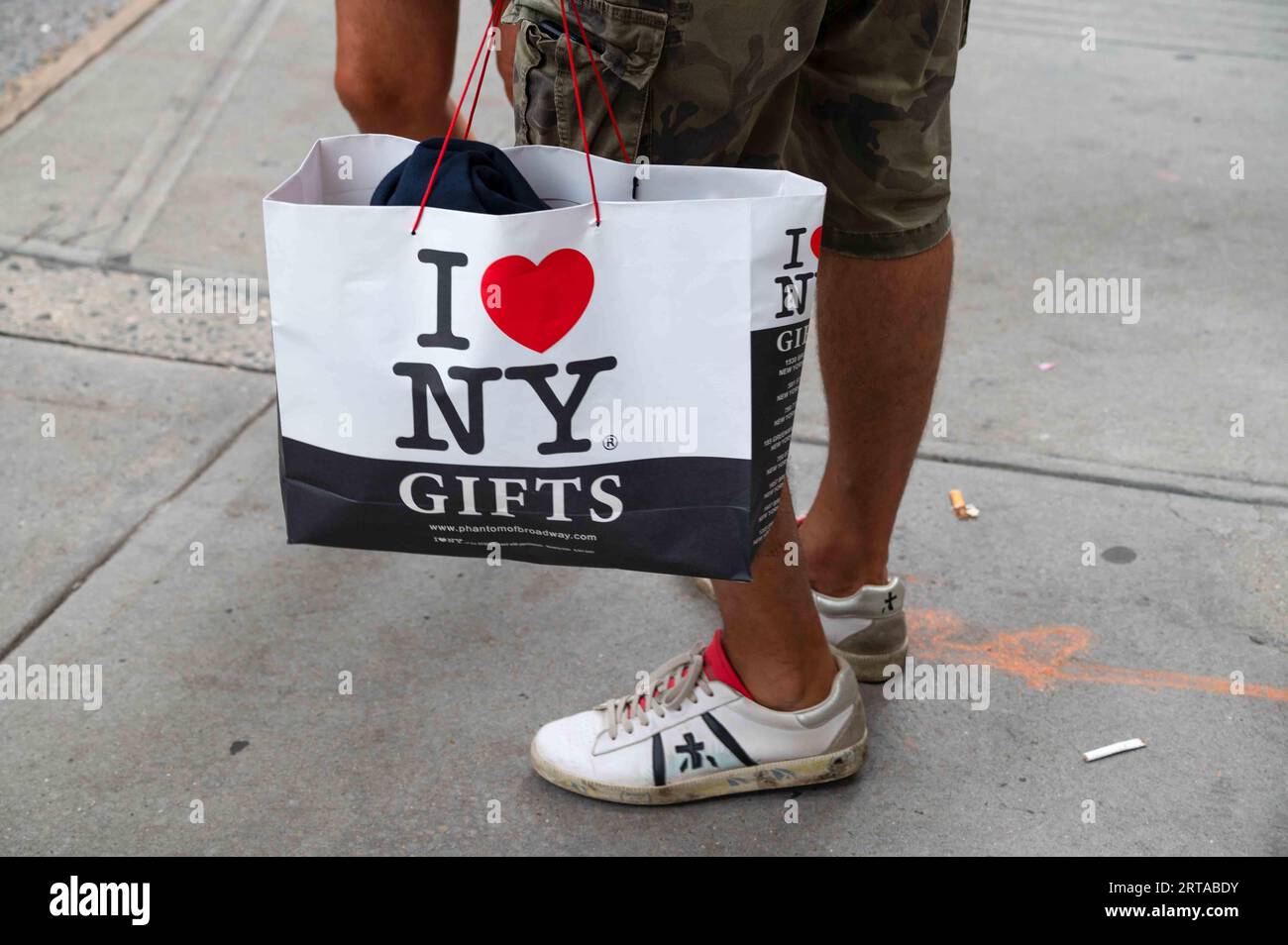 A man in a sidewalk wearing sneakers with I heart NY souvenir gift bag Stock Photo