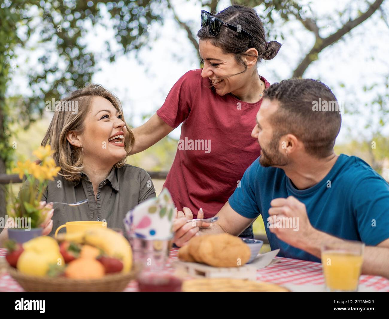 Three caucasian friends enjoy an outdoor brunch. A seated woman and a young man have a spread of food before them, while a brunette woman stands, embr Stock Photo