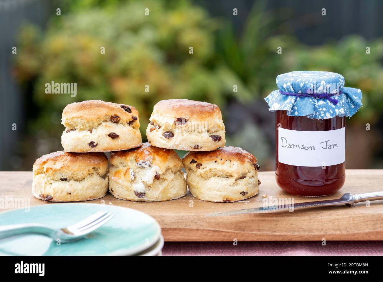 A freshly baked batch of fruit scones displayed on a serving board with a jar of homemade Damson Jam. All served to an outdoor table Stock Photo