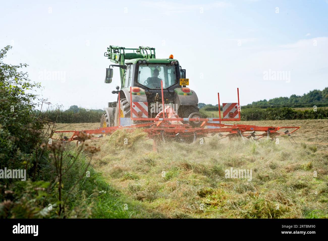 A farm worker driving a tractor across a field of freshly cut grass.The tractor has a tedding attachment which is tedding the cut grass for hay making Stock Photo