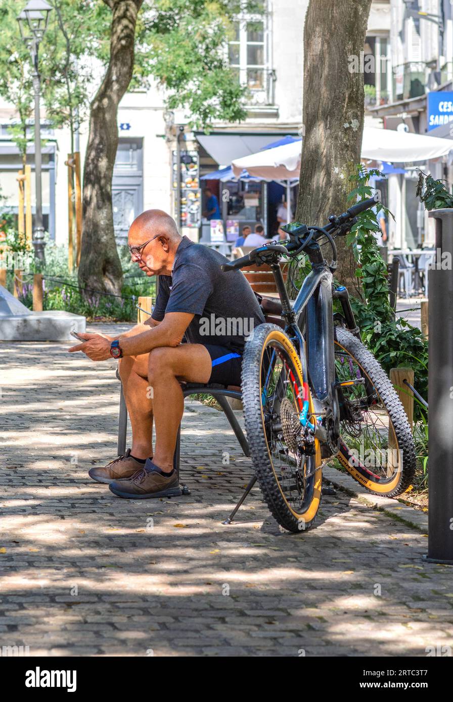 Older man with cross bike sitting on city center bench studying smartphone - Tours, Indre-et-Loire (37), France. Stock Photo