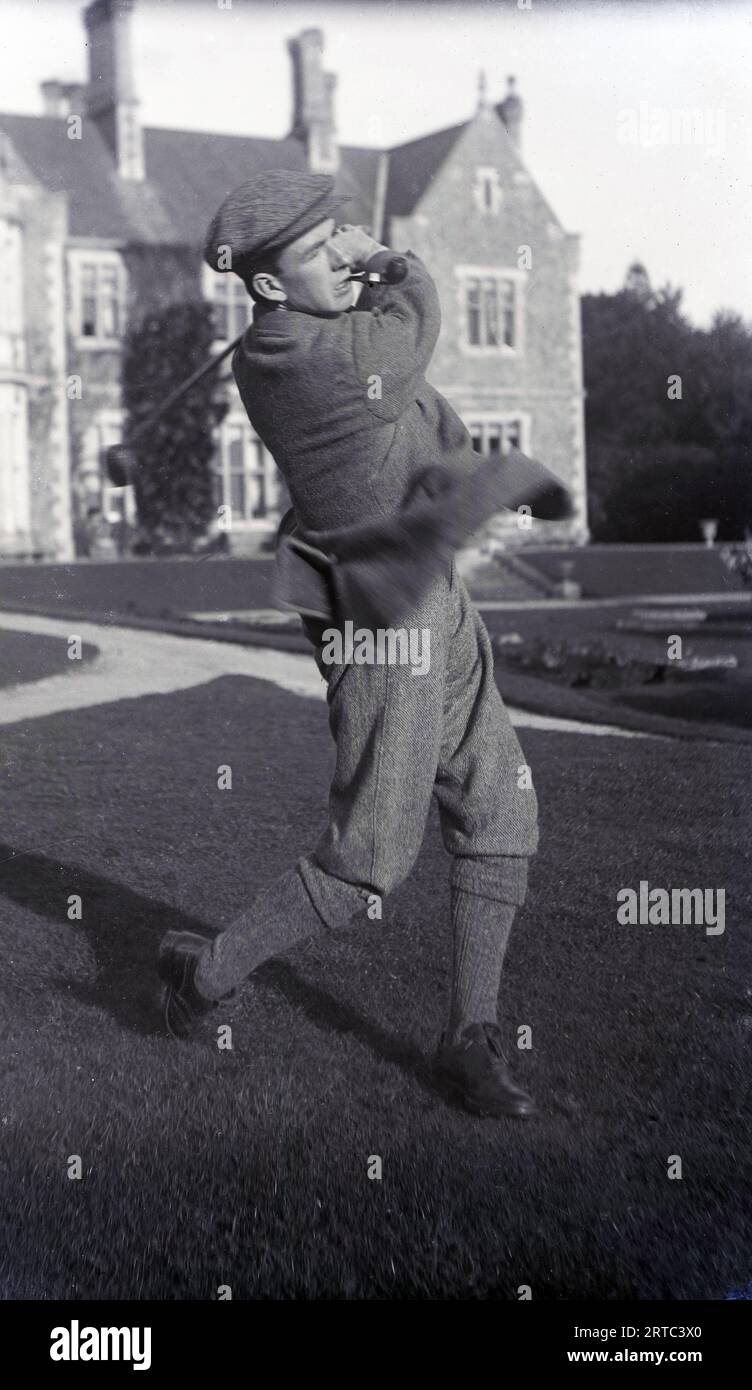 1930s, historical, outside in the grounds of a private school, a young gentleman golfer in the male dress of the era, a woollen jacket and trousers and plus fours, flat cap and pipe in mouth, swinging a golf club, England, UK. Stock Photo