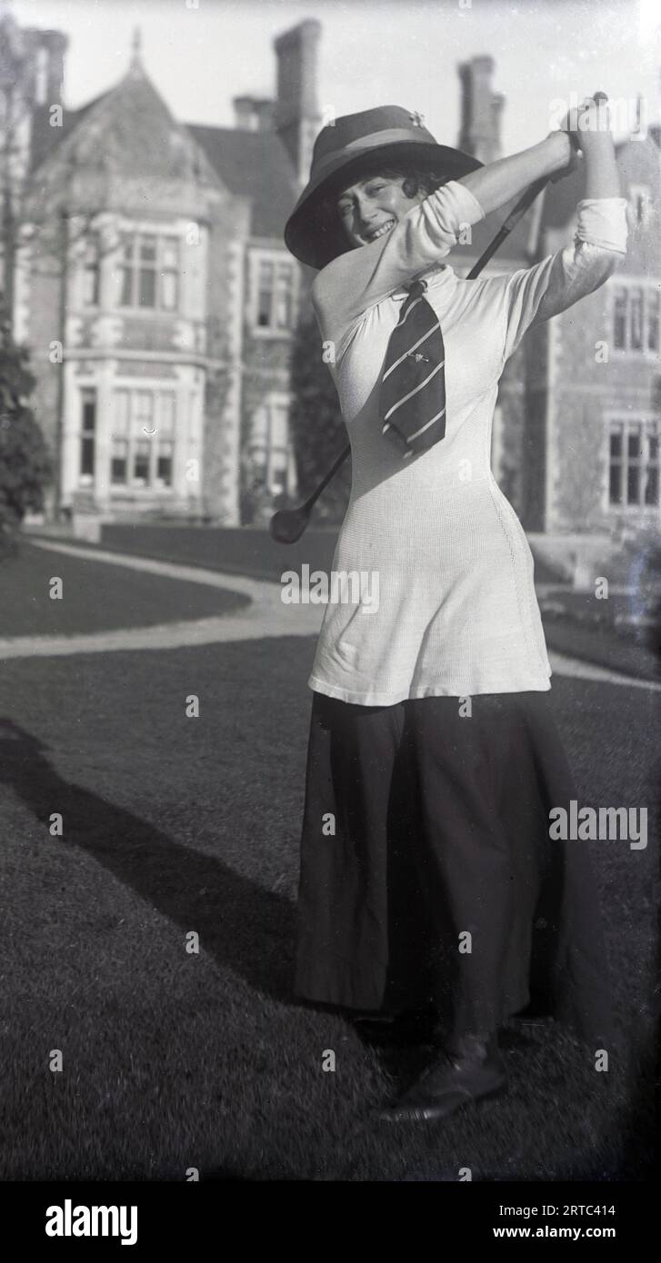 1930s, historical, in the grounds outside a private school, a young female golfer swinging a golf club, England, UK, wearing the female fashions of the day, a long shirt & tie, over an ankle length skirt and hat. Stock Photo