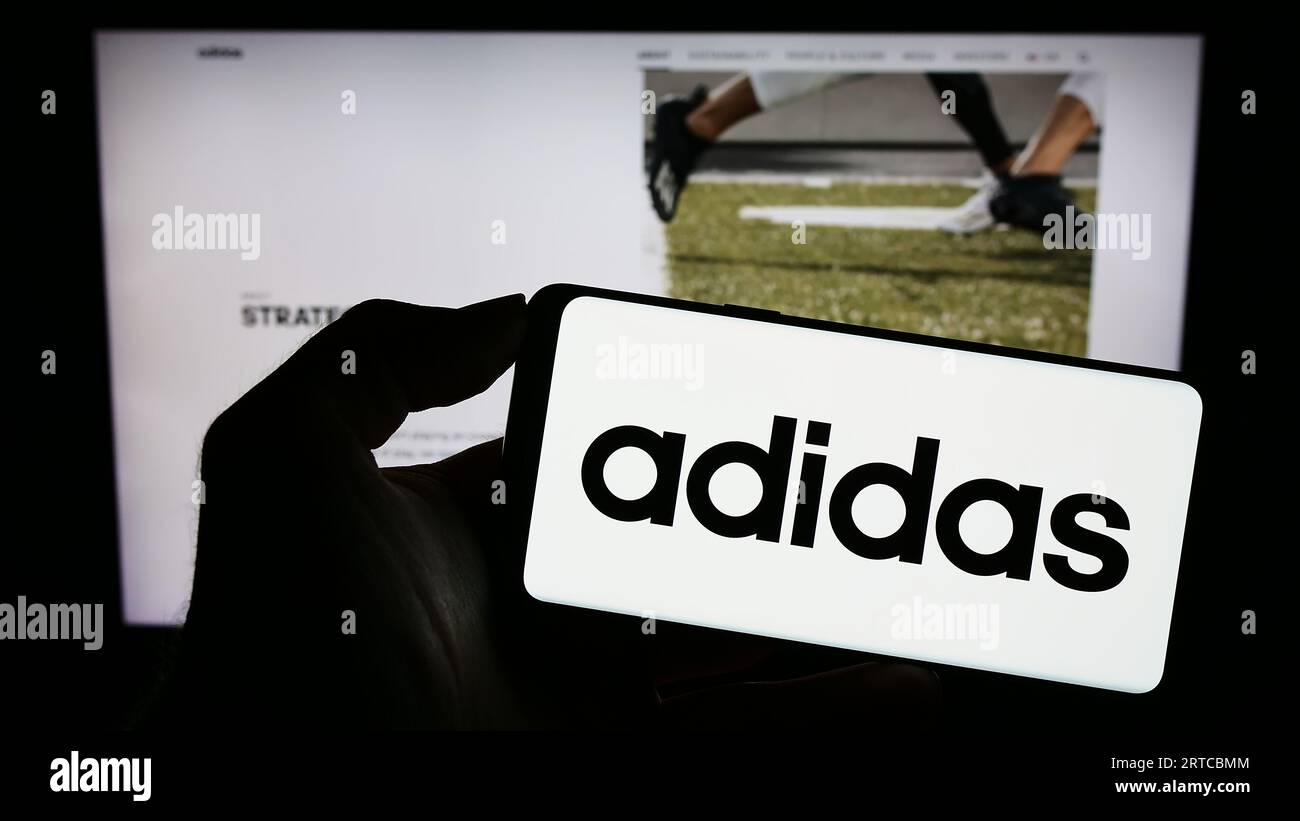 Person holding smartphone with logo of German apparel and footwear company Adidas AG on screen in front of website. Focus on phone display. Stock Photo