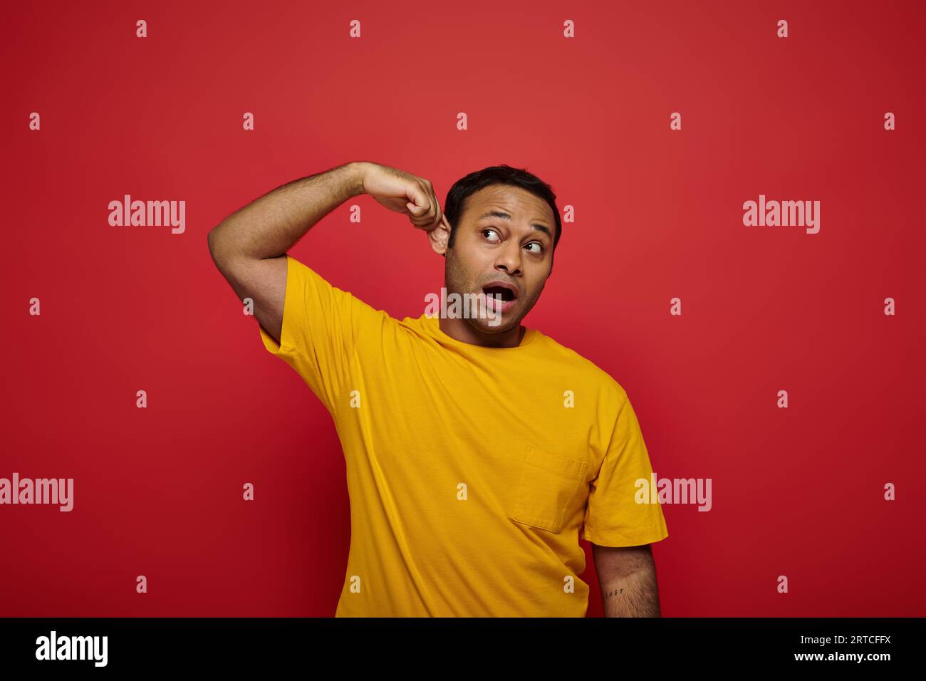 curious indian man in bright casual clothes looking away and touching ear on red background Stock Photo