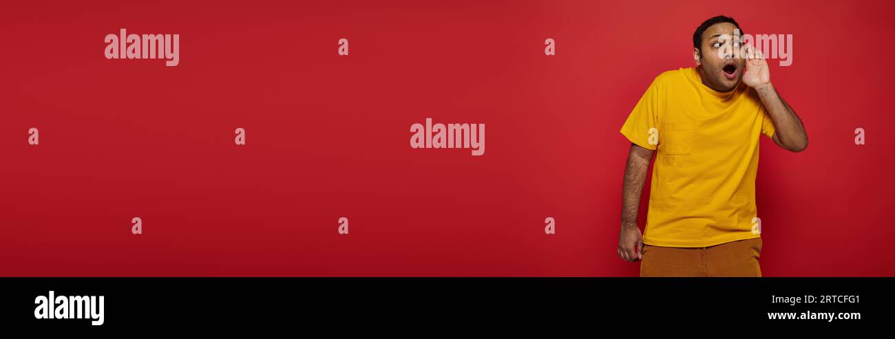 curious indian man in bright clothes looking away and listening on red background, banner Stock Photo