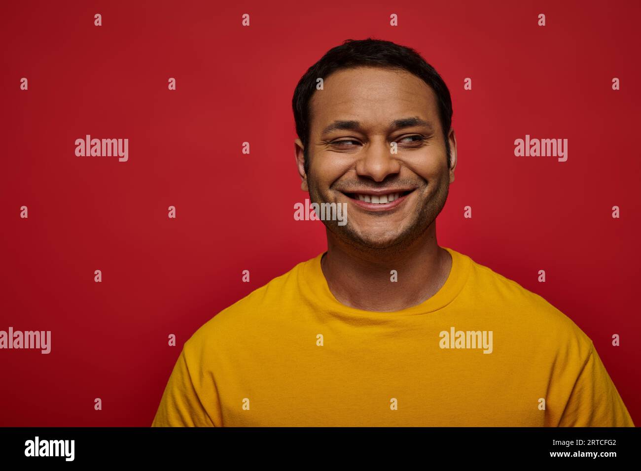 sly indian man in bright yellow clothes looking away and smiling on red background, side glance Stock Photo