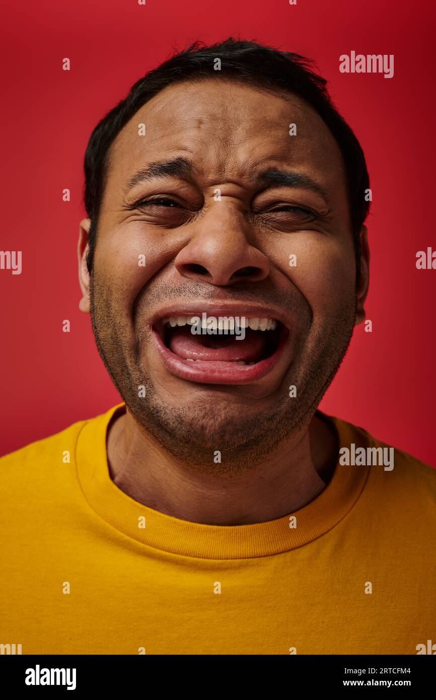 face expression, emotional indian man in yellow t-shirt crying loud on red background, open mouth Stock Photo