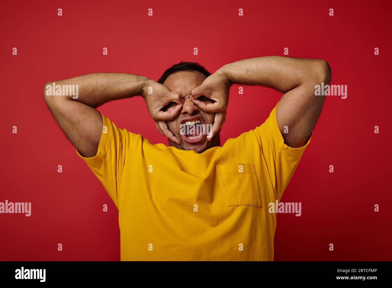 emotional indian man in yellow t-shirt screaming and gesturing on red backdrop, expressive face Stock Photo