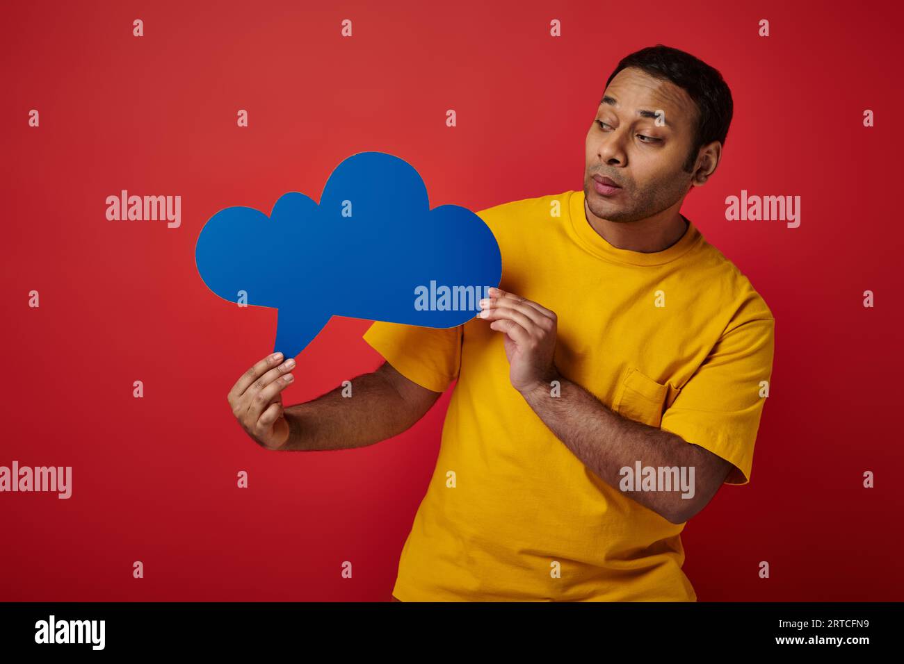 indian man in yellow t-shirt looking at blank thought bubble on red backdrop, upset face Stock Photo