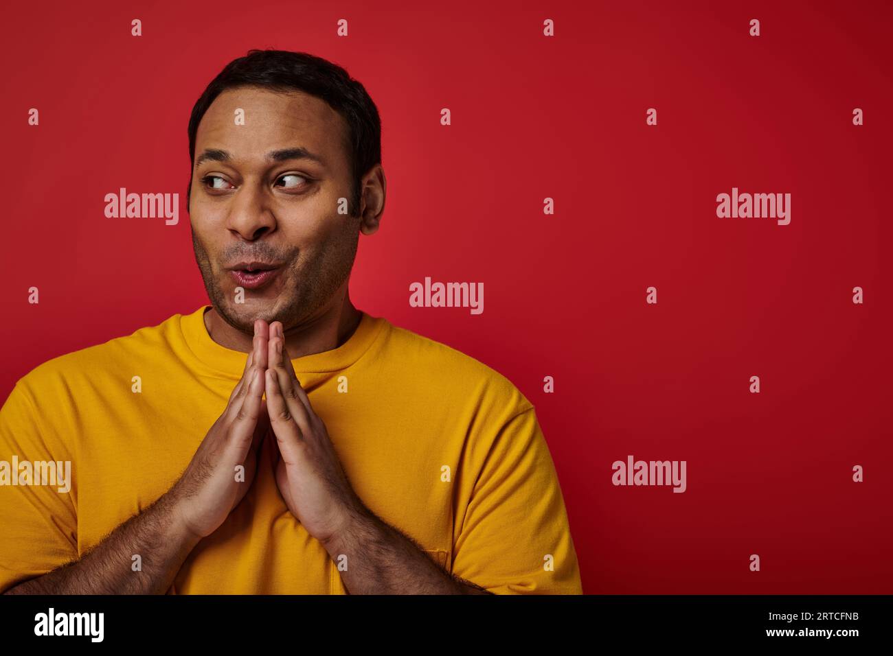 cunning indian man in yellow t-shirt clasping hands and planning something on red backdrop, sly face Stock Photo