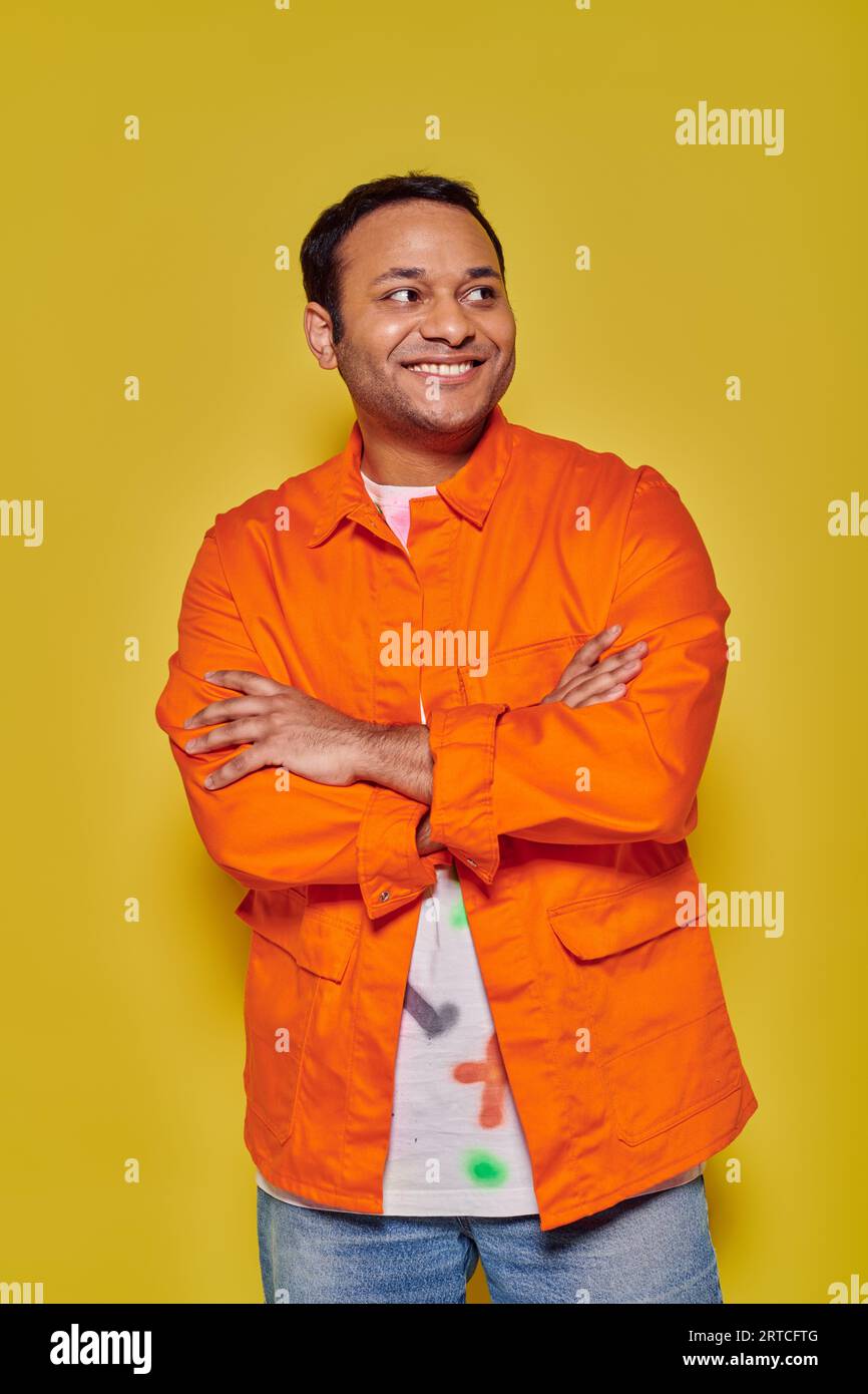 portrait of positive indian man in orange jacket standing with folded arms on yellow backdrop Stock Photo
