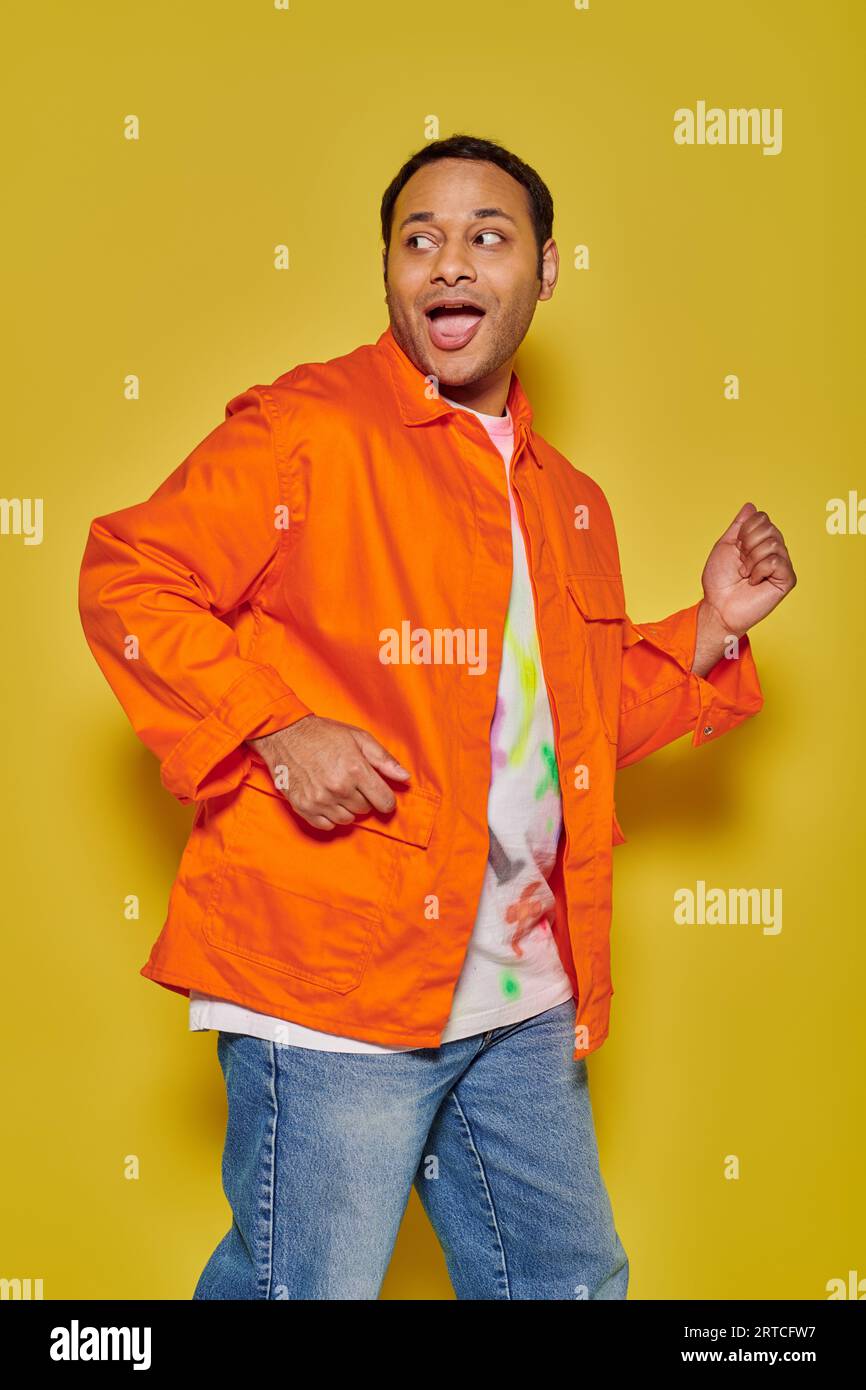 portrait of amazed indian man in orange jacket running away and looking back on yellow backdrop Stock Photo