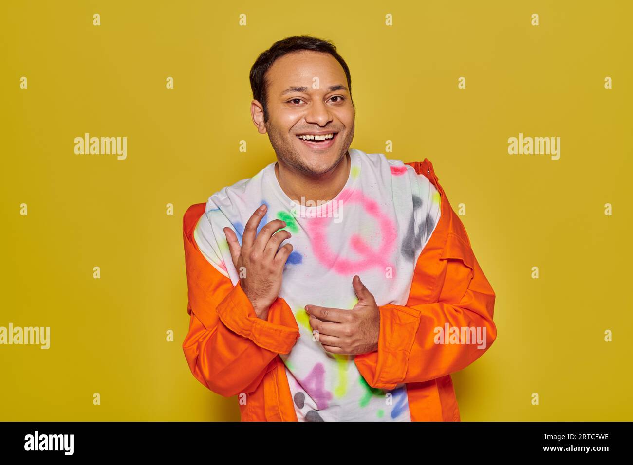 gleeful indian man in orange jacket and diy t-shirt smiling and looking at camera on yellow backdrop Stock Photo