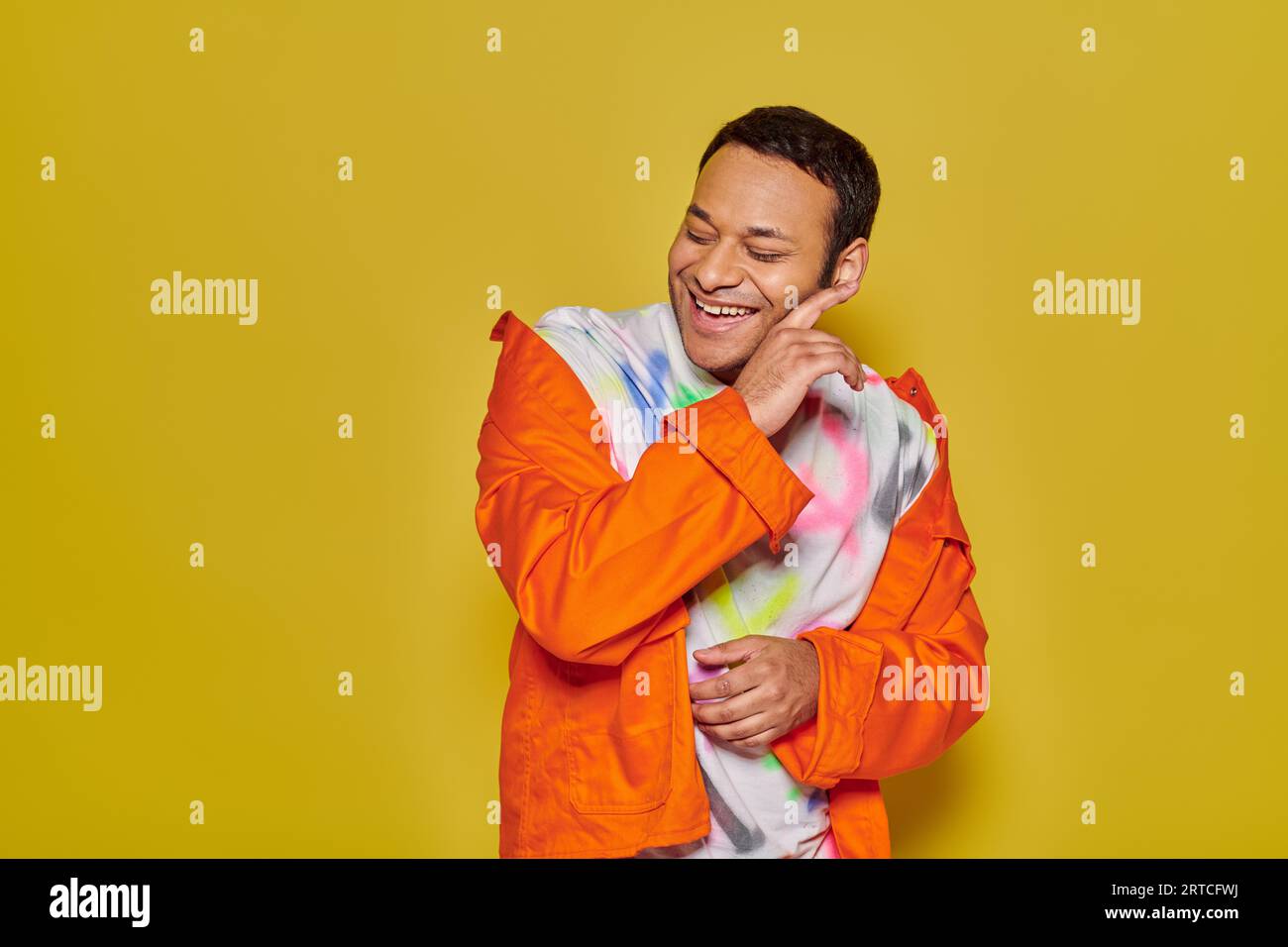 gleeful indian man in orange jacket and diy t-shirt smiling with closed eyes on yellow backdrop Stock Photo