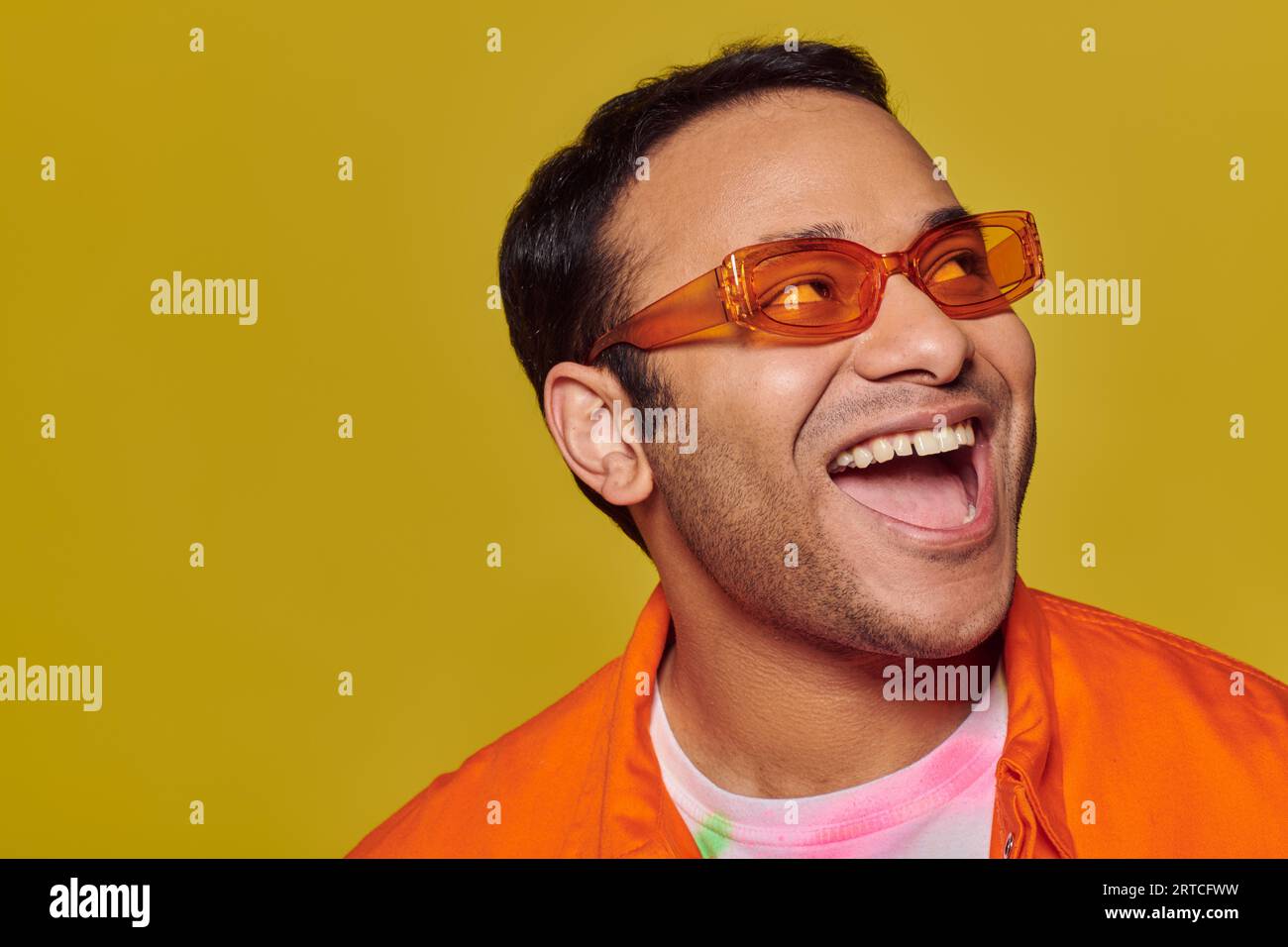 positive indian man in orange sunglasses looking away and smiling on yellow background, side glance Stock Photo