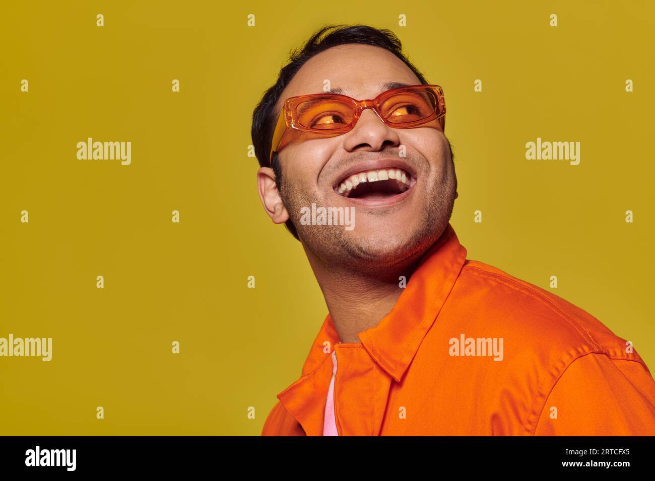 amazed indian man in orange sunglasses looking away and smiling on yellow background, side glance Stock Photo