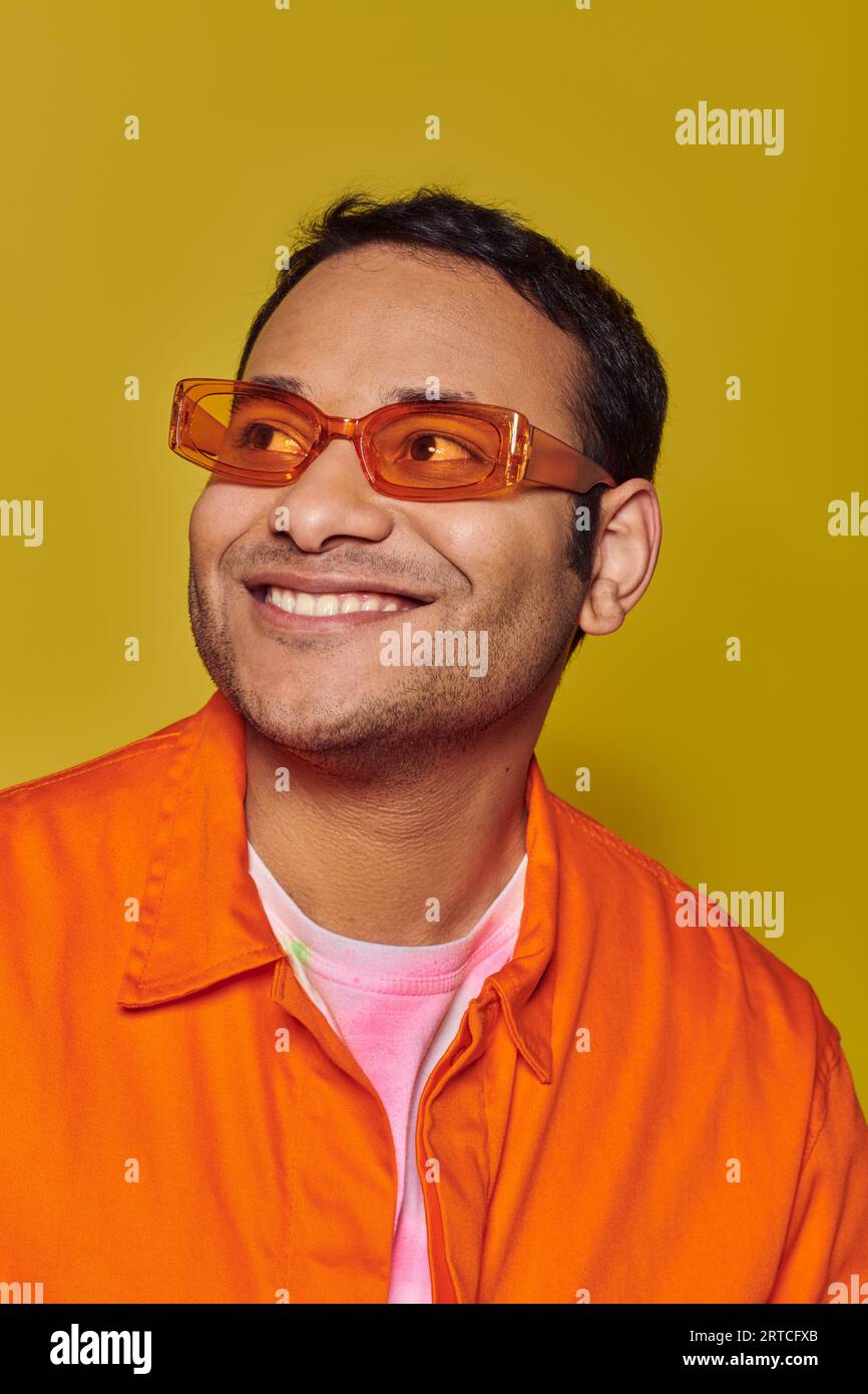 happy indian man in orange sunglasses looking away while smiling on yellow background, side glance Stock Photo