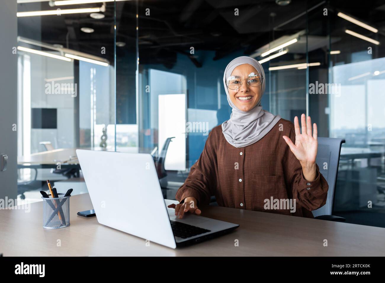 Portrait of a young Muslim woman in hijab sitting at a desk in the office, working and studying on a laptop, looking and waving at the camera. Stock Photo
