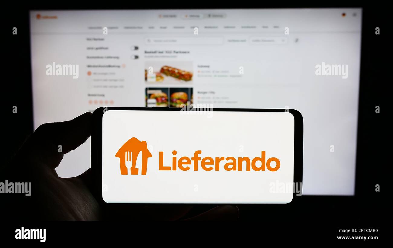 Person holding mobile phone with logo of German food delivery company Lieferando on screen in front of business web page. Focus on phone display. Stock Photo