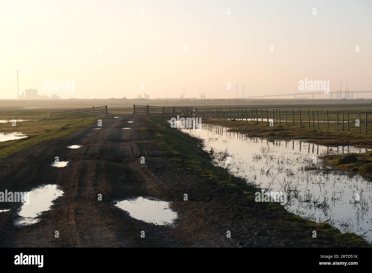 Wet farm track and gate with Elmley Cement Works and Kingsferry Bridge in the background, Elmley, Isle of Sheppey, Kent, England Stock Photo