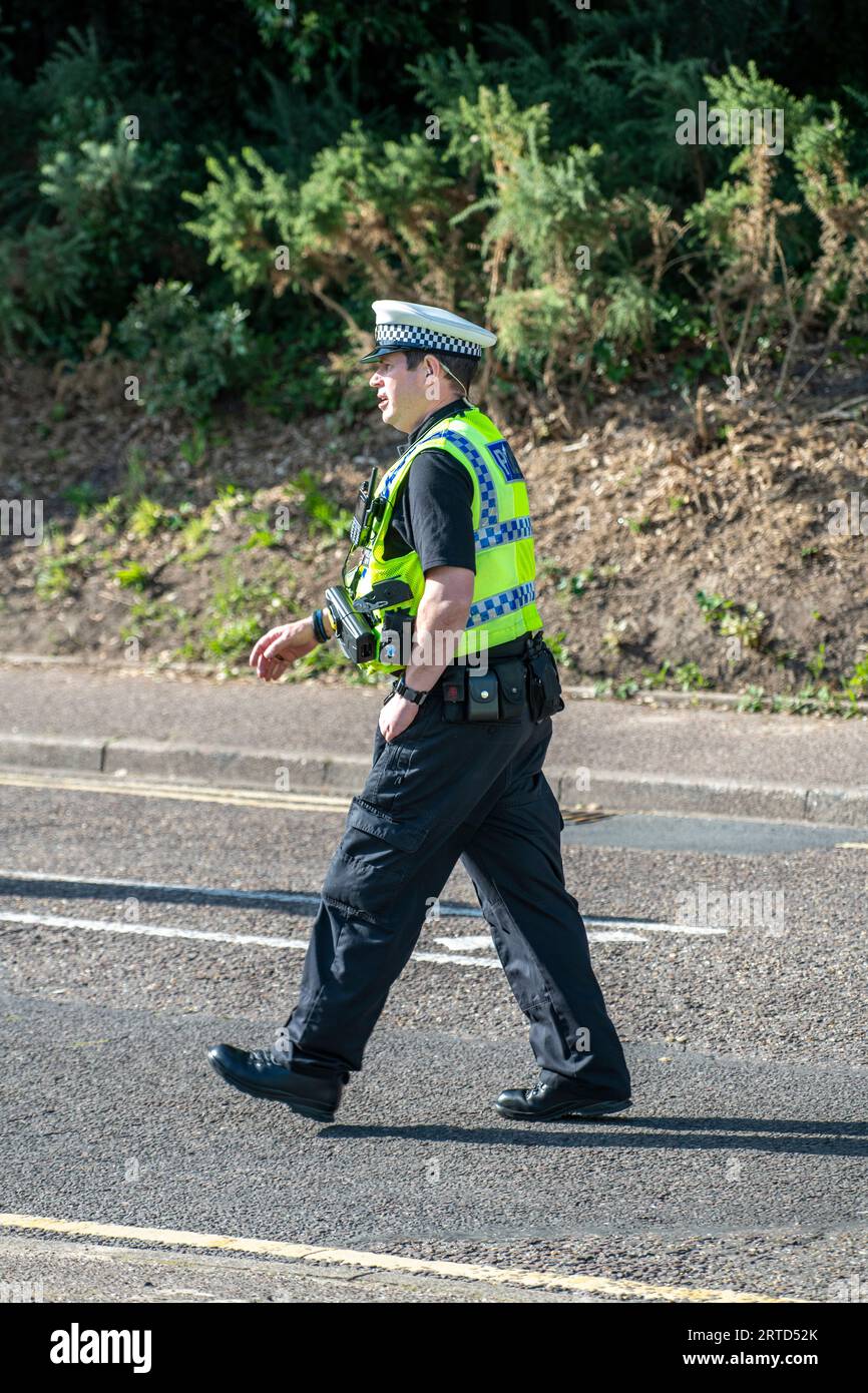 police officer wearing a cap and a high visibility jacket walking along a road on a sunny day. Stock Photo