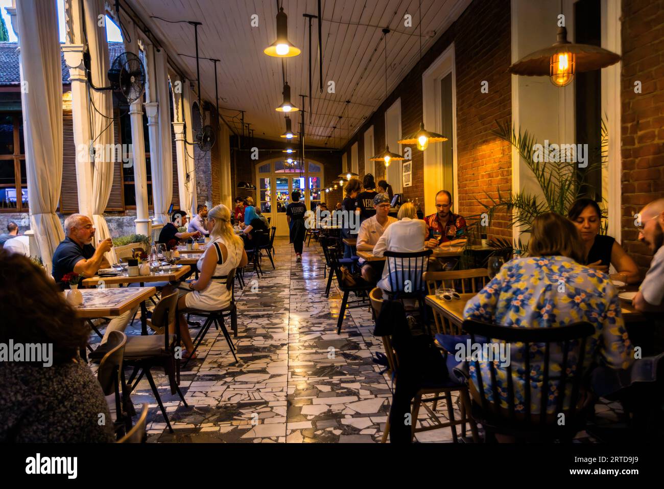 Evening scene on the covered veranda of the Kikodze Coffee & Cocktail Bar in Vardisubani, Tbilisi: guests sit beneath hanging lamps on ornamental concrete tiles, whilst the exposed clinker brick wall and curtains between white columns frame the long restaurant corridor. The bar forms part of a historic complex of buildings in the Vardisubani district. Vasil Petriashvili Street, Vardisubani, Vera, Mtatsminda District, Tbilisi, 0108, Georgia Stock Photo