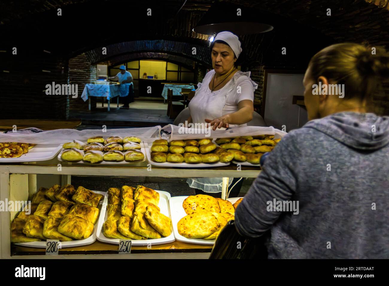The best baked goods in Tbilisi can be found in the Sophiko Chiaureli Garden. A woman in a white dress and headscarf stands behind a counter and offers baked goods. Numerous baked goods lie on trays in the display in front of a person. Another person can be seen in the background. Tbilisi, Georgia. Stock Photo