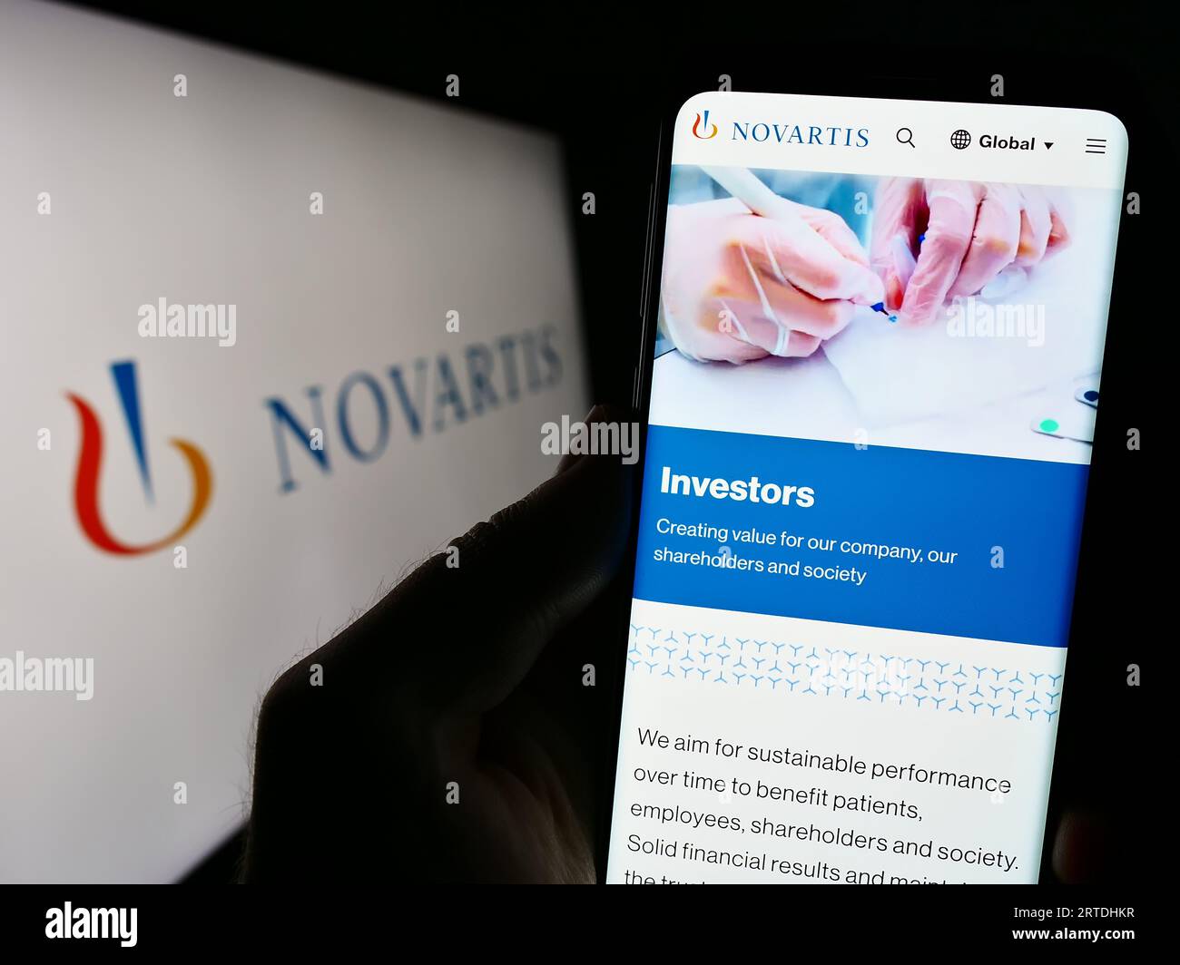 Person holding cellphone with webpage of Swiss pharmaceutical company Novartis AG on screen in front of logo. Focus on center of phone display. Stock Photo