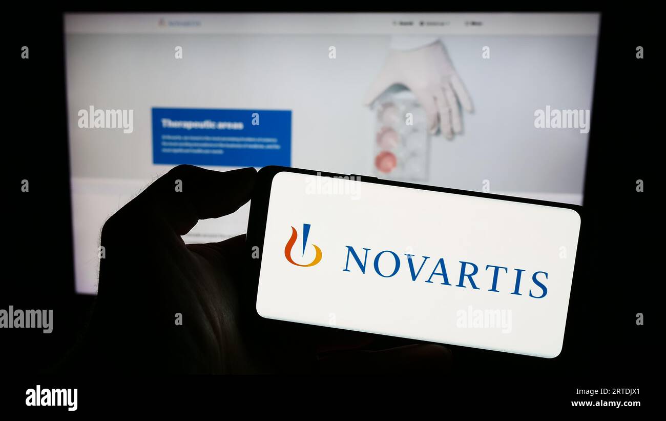 Person holding smartphone with logo of Swiss pharmaceutical company Novartis AG on screen in front of website. Focus on phone display. Stock Photo